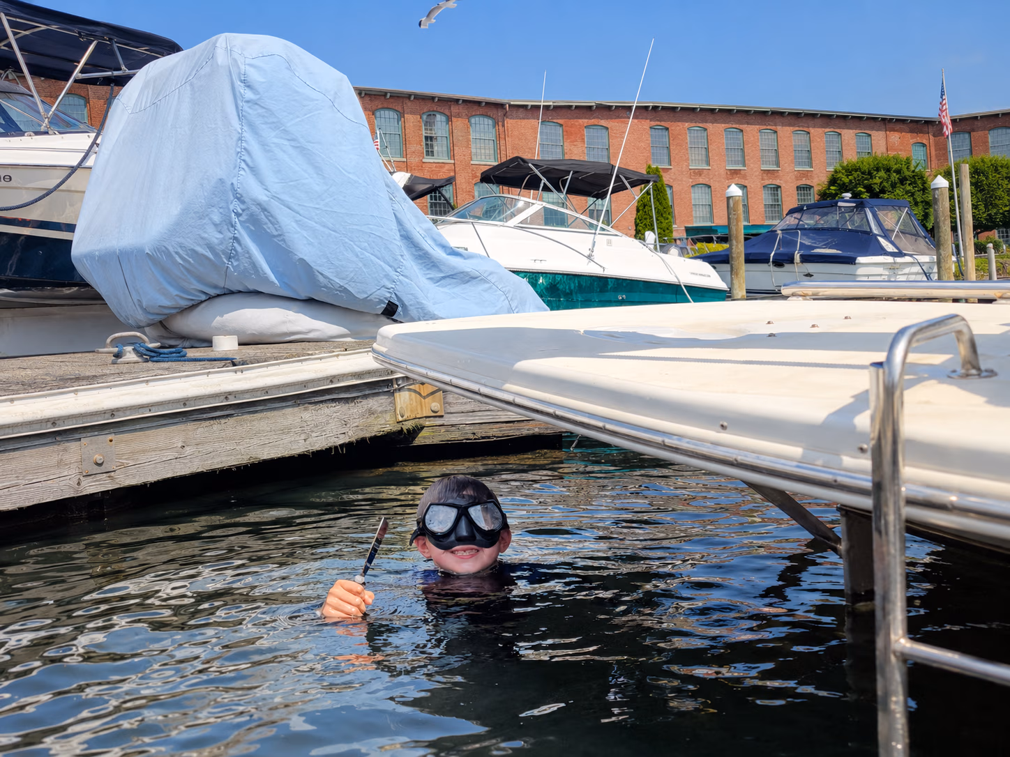 Child wearing goggles smiling in water near dock and boats, holding a marine tool or knife.
