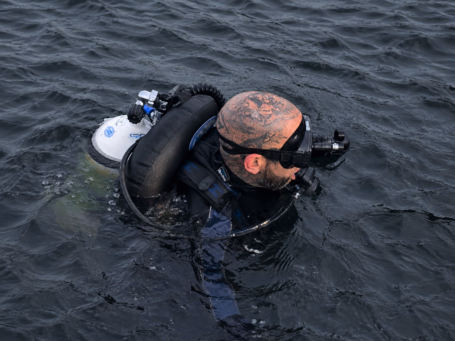 Scuba diver with tattooed shaved head, wearing mask and breathing apparatus, floating in dark water.