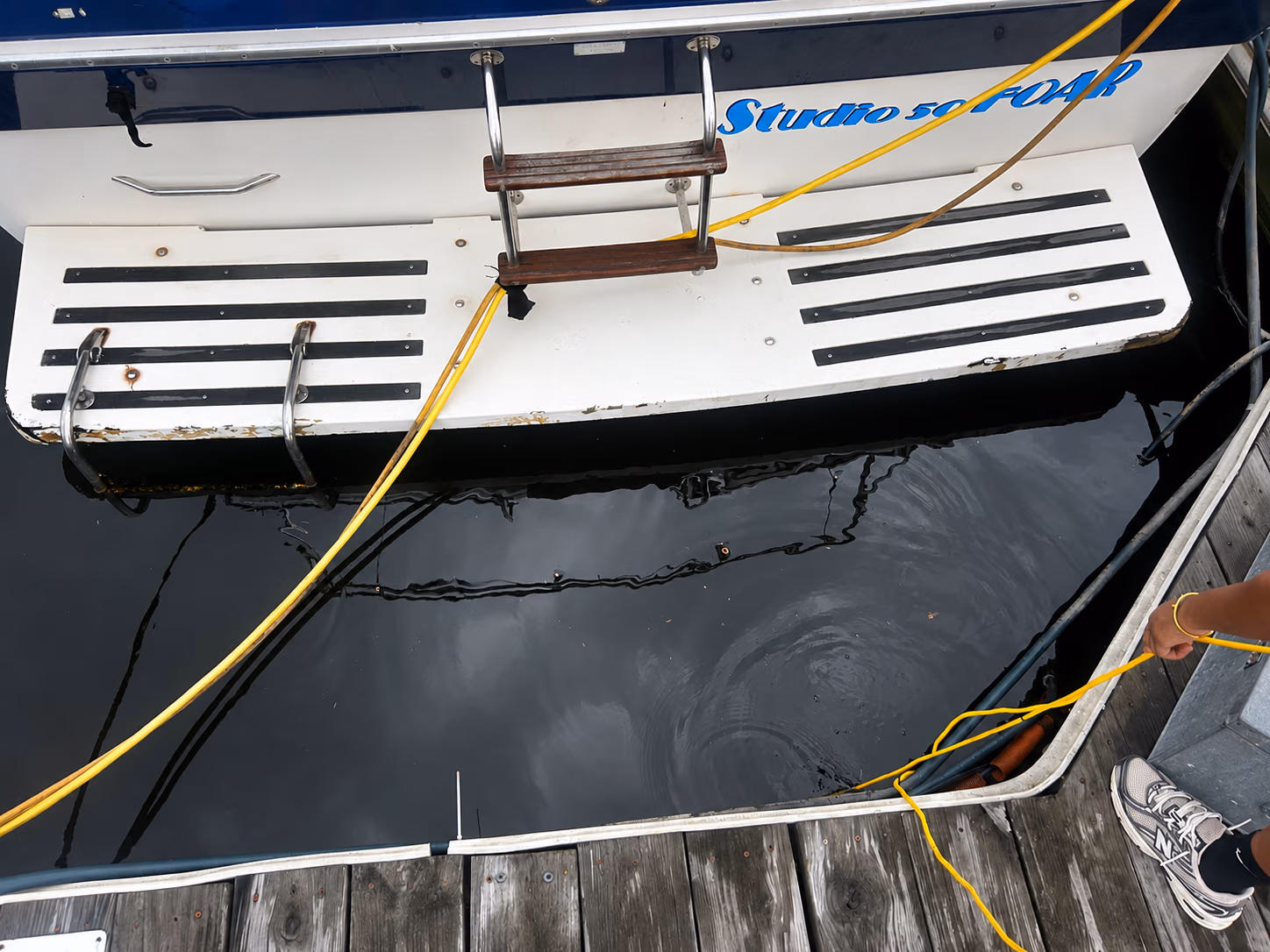 Boat named Studio 50 FOAR docked with yellow cables and a person's hand holding a cable on a wooden dock.