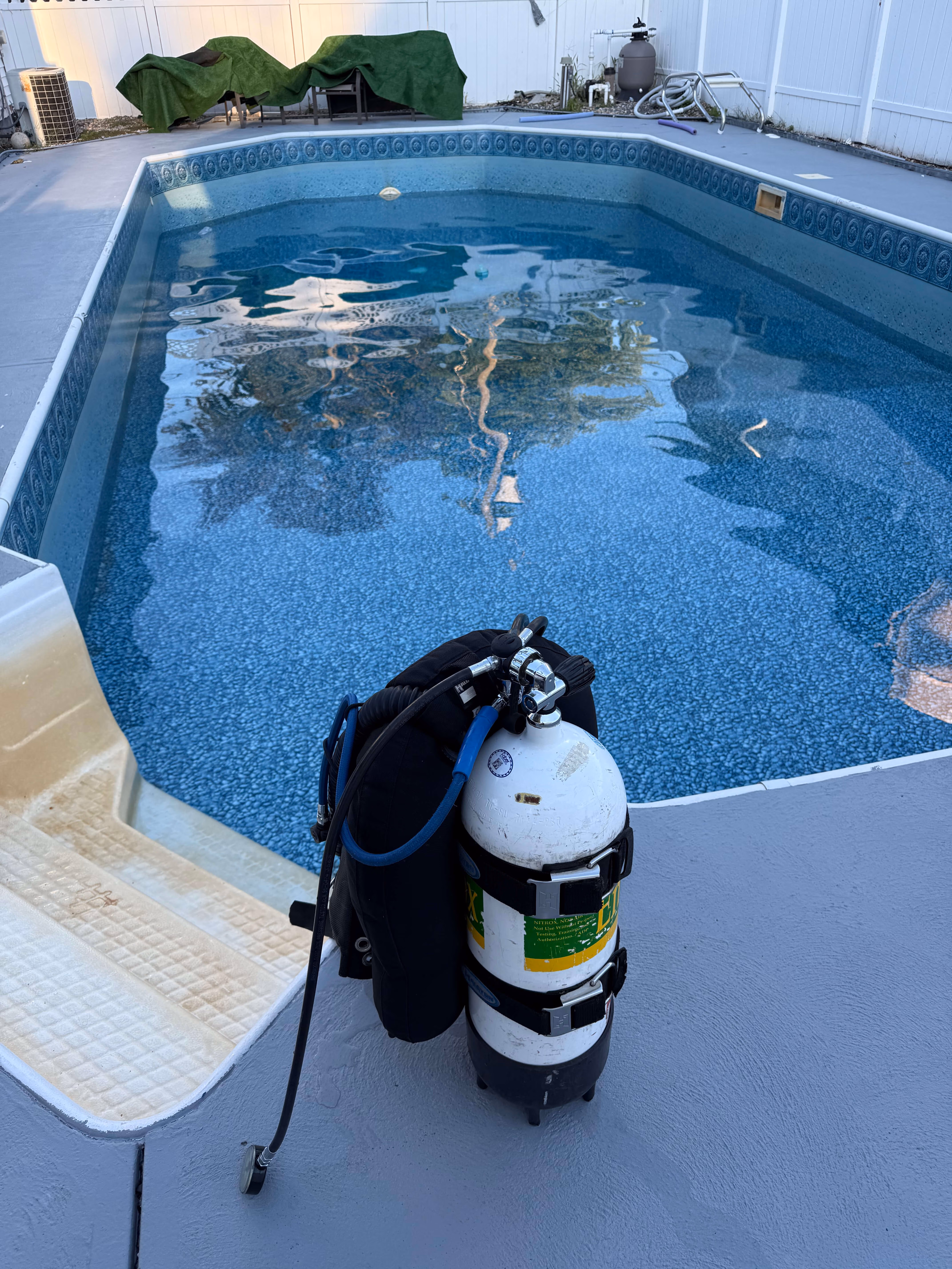 Scuba diving tank with hoses standing on pool deck next to in-ground pool with steps.