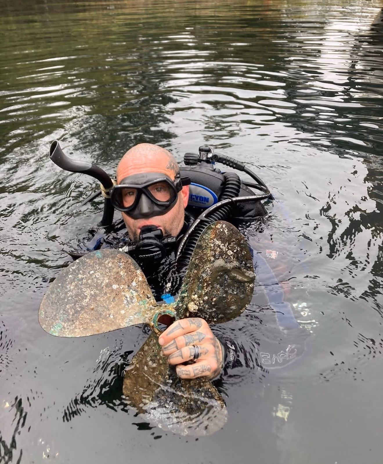 Scuba diver holding a large, barnacle-covered boat propeller while floating in water.