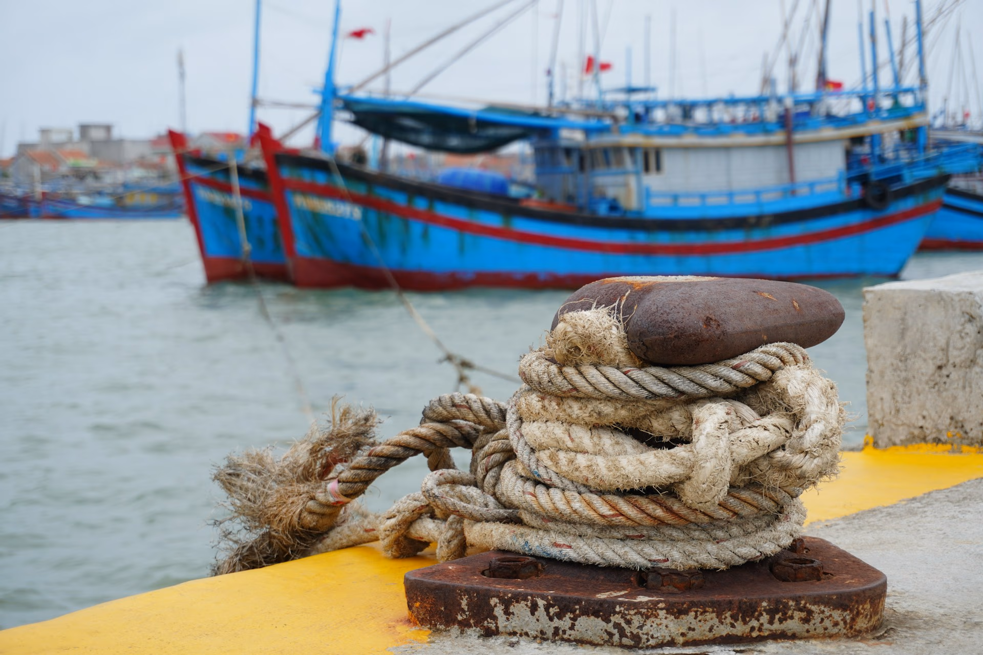 Rusty metal cleat with coiled rope on a dock, blue fishing boats moored in background.