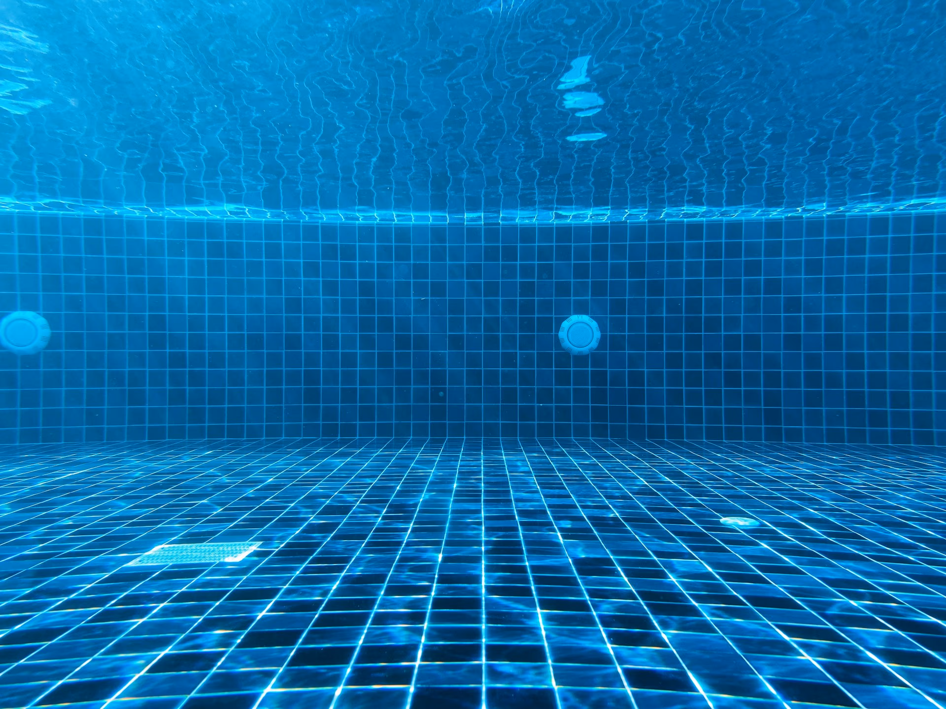 Underwater view of a tiled pool floor and walls with two circular pool fittings visible.