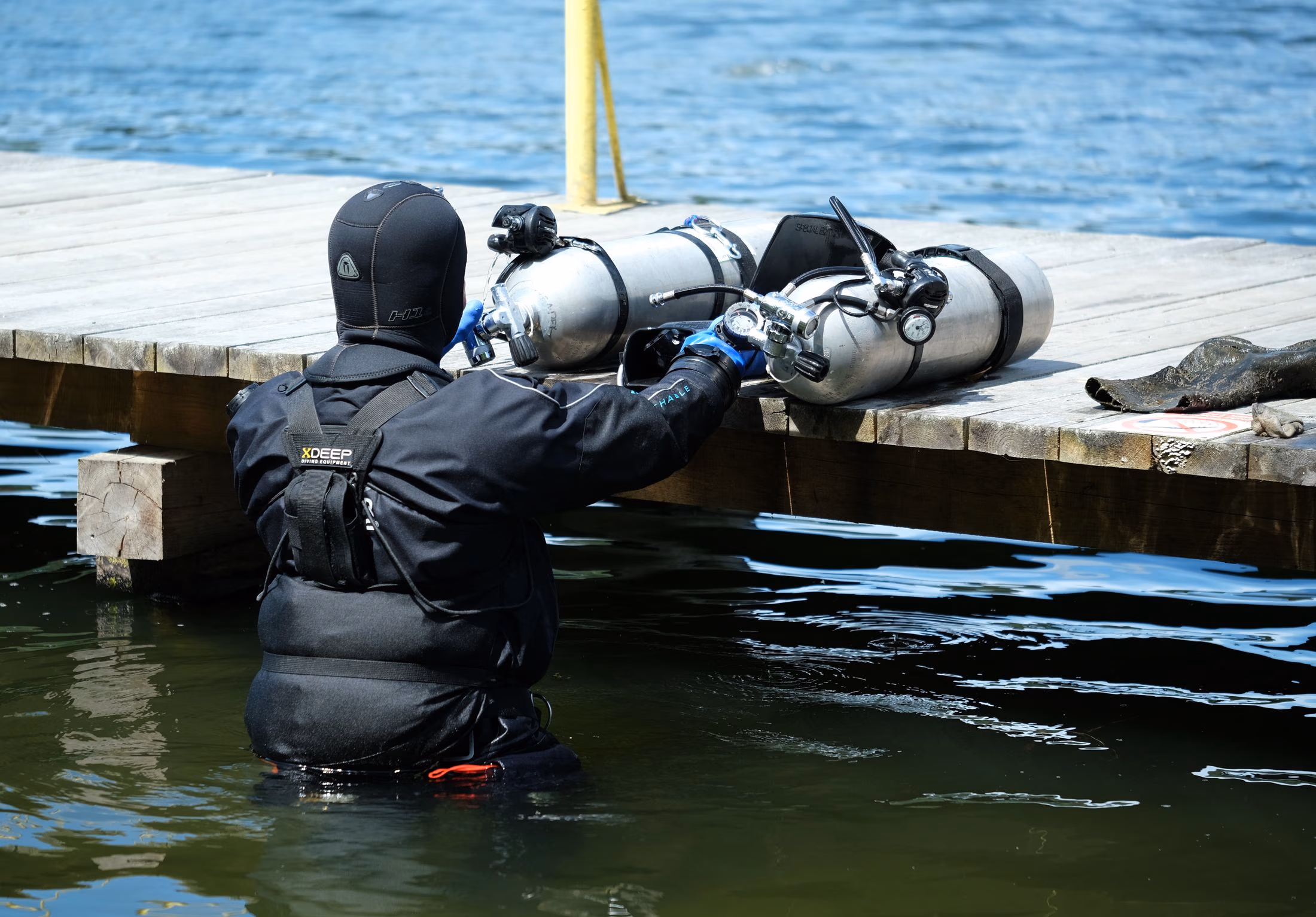 Diver in wetsuit standing in water by a dock, handling two scuba tanks on the dock.